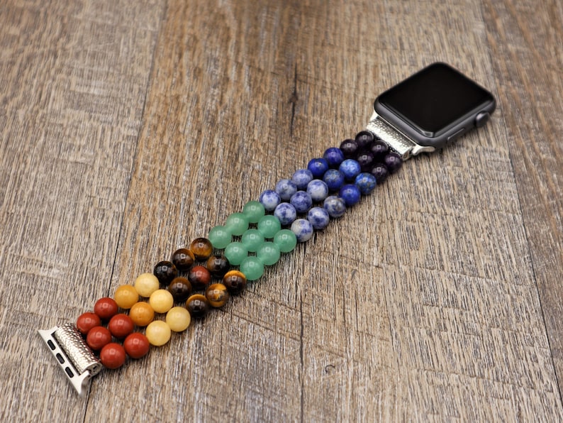 Op de afbeelding: Een kleurrijk kralenbandje voor de Apple Watch met rode, oranje, gele, groene, blauwe en paarse kralen. Het bandje is gemaakt van natuurlijke stenen en is ontworpen om te passen op een standaard Apple Watch.
