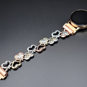 May include: A rose gold smartwatch band with a heart-shaped design. The band is made of metal and has a silver, gold, and rose gold finish. The band is designed to fit a smartwatch with a standard 20mm band size.