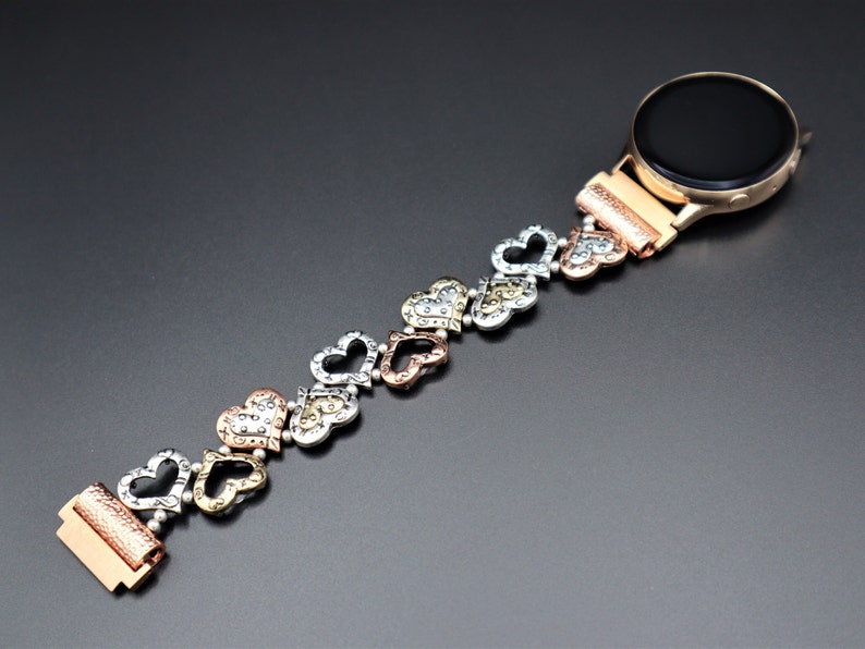 May include: A rose gold smartwatch band with a heart-shaped design. The band is made of metal and has a silver, gold, and rose gold finish. The band is designed to fit a smartwatch with a standard 20mm band size.
