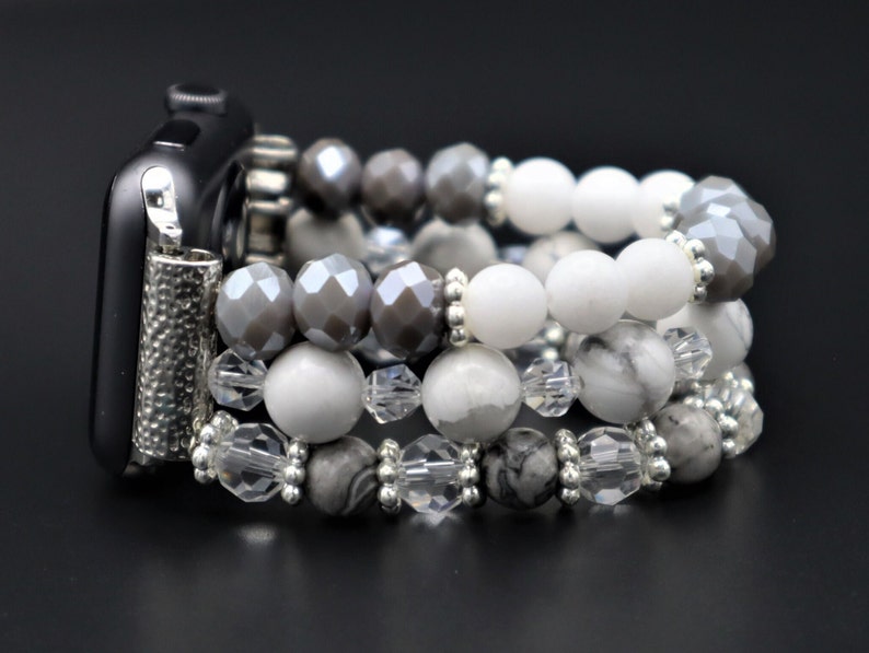 May include: A silver and white beaded watch band with a black watch face. The band is made of multiple strands of beads, including white, grey, and clear beads.