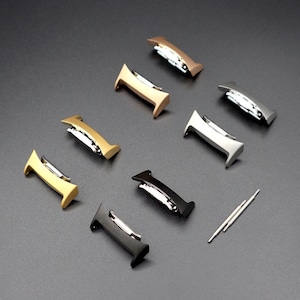 May include: A set of seven watch band spring bars in various metallic finishes, including gold, silver, and black. The spring bars are arranged in a row on a gray surface, with two small metal tools for installing the spring bars.