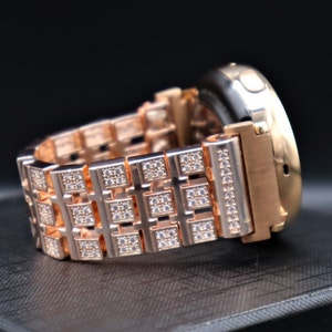 May include: Rose gold watch with a band featuring square links adorned with small, sparkling crystals. The watch case is also rose gold, with a black watch face. The band is made of metal and has a secure clasp.