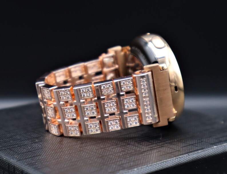 May include: Rose gold watch with a band featuring square links adorned with small, sparkling crystals. The watch case is also rose gold, with a black watch face. The band is made of metal and has a secure clasp.