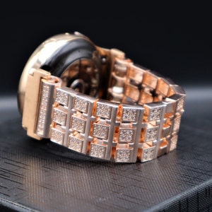 May include: Rose gold watch strap with a square link design, adorned with small, sparkling crystals. The watch face is black, and the strap is on a textured black surface.