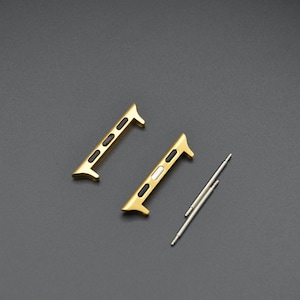 May include: Two gold-colored watch band adapters with black and white accents, and two silver spring bars. The adapters have a rectangular shape with rounded edges and are designed to connect a watch band to a watch case.