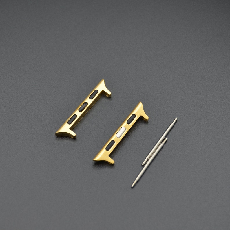 May include: Two gold-colored watch band adapters with black and white accents, and two silver spring bars. The adapters have a rectangular shape with rounded edges and are designed to connect a watch band to a watch case.