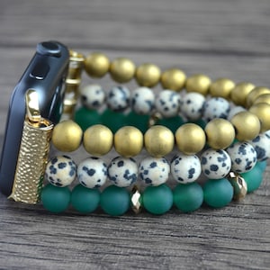 Puede incluir: Una pulsera de reloj Apple de cuentas doradas, verdes y blancas. La pulsera está hecha de tres hileras de cuentas: doradas, verdes y blancas con manchas negras.