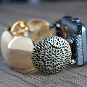 Puede incluir: Una pulsera de reloj de metal dorado y negro con un diseño texturizado. La pulsera está compuesta por una serie de piezas redondas que se superponen.