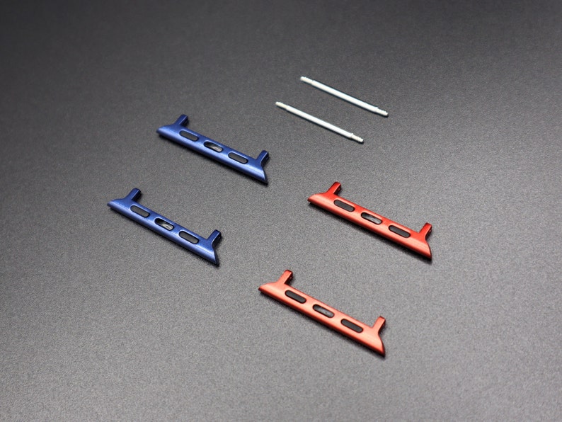 May include: An overhead shot displays watch band adapters in blue and red, along with two silver pins. The adapters have a rectangular shape with rounded edges and three small, rectangular cutouts. The background is a dark gray surface.