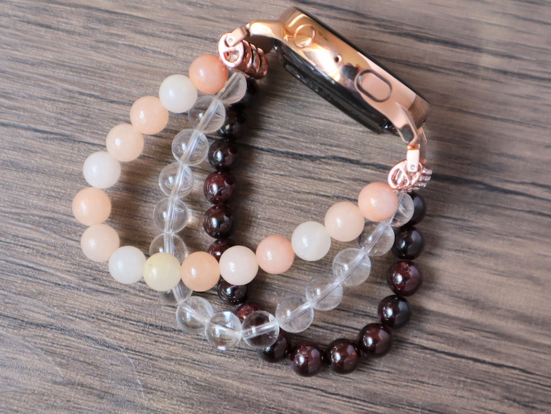 May include: A rose gold smartwatch band made of three strands of beads. The top strand is peach colored, the middle strand is clear, and the bottom strand is dark red.
