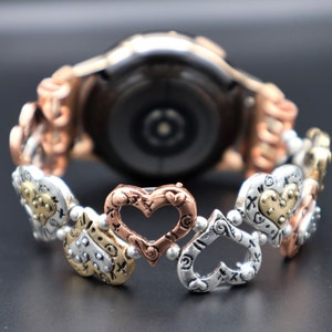 May include: A silver, gold, and rose gold watch band with a heart-shaped design. The band is made of metal and has a polished finish.