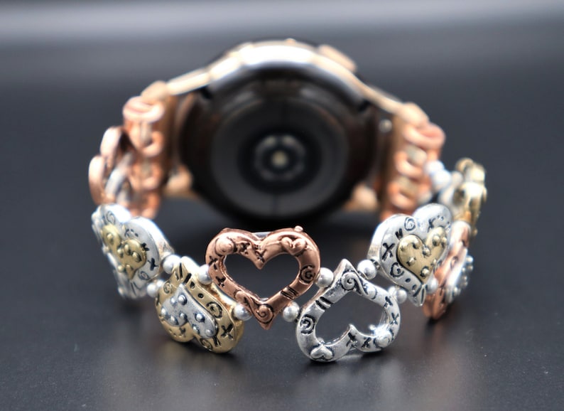 May include: A silver, gold, and rose gold watch band with a heart-shaped design. The band is made of metal and has a polished finish.