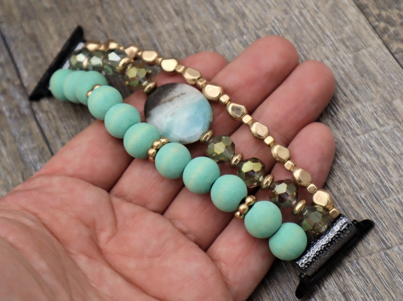May include: A multi-strand watch band with turquoise wooden beads, gold beads, and clear faceted glass beads. A large turquoise gemstone is featured in the center of the band.