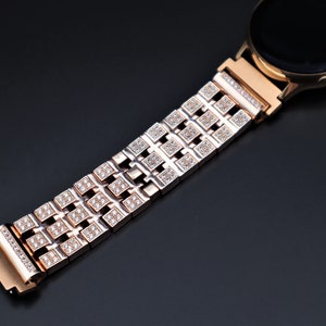 May include: Rose gold smartwatch with a matching strap. The strap has a repeating pattern of square links, each set with small, sparkling crystals. The watch face is black, and the design is elegant and modern.