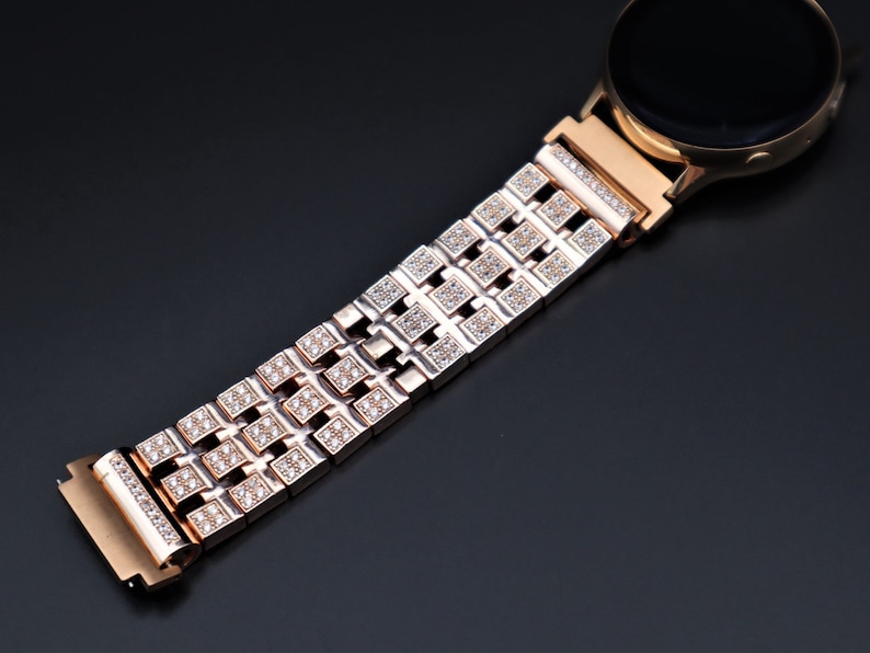 May include: Rose gold smartwatch with a matching strap. The strap has a repeating pattern of square links, each set with small, sparkling crystals. The watch face is black, and the design is elegant and modern.