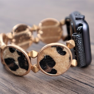 Puede incluir: Una correa de reloj dorada con estampado de leopardo y una esfera negra. La correa está hecha de metal y tiene un diseño redondo y circular.