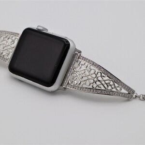 May include: A silver Apple Watch with a decorative, intricate, silver band with clear stones. The band is a delicate, openwork design.