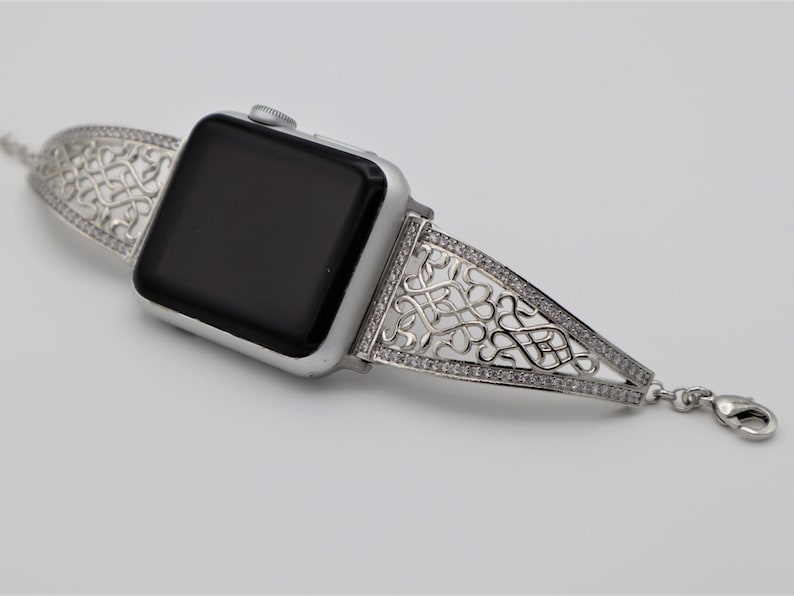 May include: A silver Apple Watch with a decorative, intricate, silver band with clear stones. The band is a delicate, openwork design.