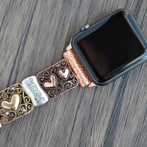 Puede incluir: Un Apple Watch con una correa decorativa con dise&ntilde;os de corazones y volutas. La correa est&aacute; hecha de varios tonos met&aacute;licos, incluyendo plata, cobre y oro. La palabra "Faith" est&aacute; inscrita en uno de los eslabones.