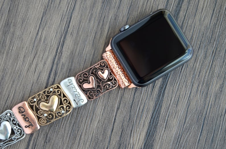 Puede incluir: Un Apple Watch con una correa decorativa con dise&ntilde;os de corazones y volutas. La correa est&aacute; hecha de varios tonos met&aacute;licos, incluyendo plata, cobre y oro. La palabra "Faith" est&aacute; inscrita en uno de los eslabones.