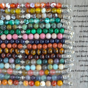 May include: A close-up of 15 strands of gemstone beads, each with a silver clasp. The beads are arranged in rows, with each row representing a different type of gemstone. The gemstones include fire agate, olive agate, coffee agate, green agate, African turquoise, malachite, rhodonite, blue-sand stone, golden-sand stone, yellow tiger's eye, red tiger's eye, Indian agate, amazonite, volcano cherry quartz, fluorite, and mookaite.