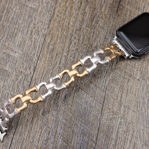 May include: An Apple Watch with a silver, gold, and silver chain-link band. The band is made of metal and has a decorative design. The watch face is black and rectangular. The watch is displayed on a wooden surface.