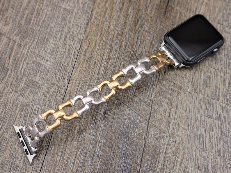 May include: An Apple Watch with a silver, gold, and silver chain-link band. The band is made of metal and has a decorative design. The watch face is black and rectangular. The watch is displayed on a wooden surface.