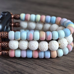 Puede incluir: Una pulsera de reloj de varias capas hecha de cuentas de colores pastel y cuentas de roca de lava blanca. La pulsera está asegurada con cierres de metal de tono cobre.