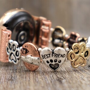 Puede incluir: Una correa de reloj en oro rosa y plata, adornada con abalorios. Los abalorios incluyen huellas de patas, corazones y un coraz&oacute;n con la inscripci&oacute;n "BEST FRIEND". La correa descansa sobre una superficie de madera.