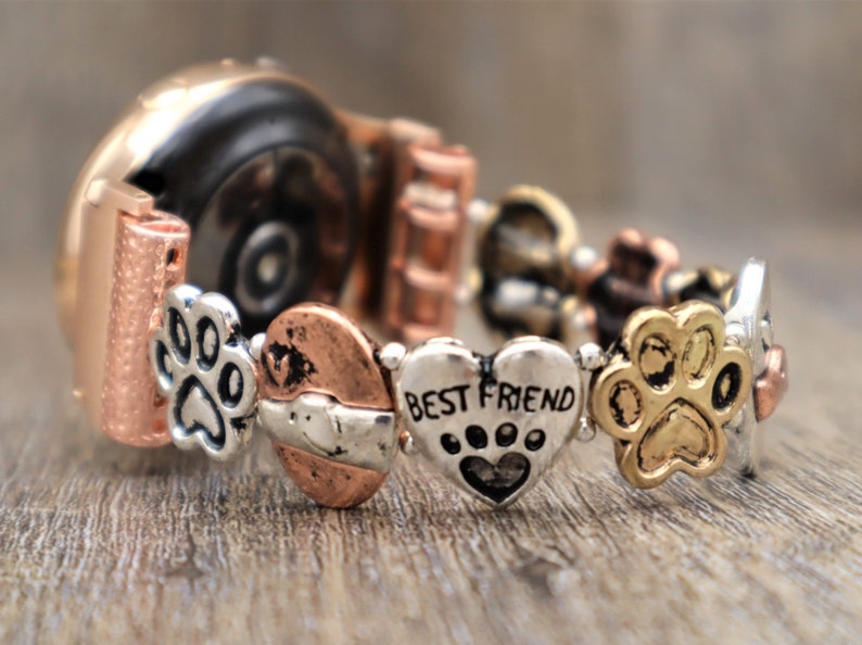 Puede incluir: Una correa de reloj en oro rosa y plata, adornada con abalorios. Los abalorios incluyen huellas de patas, corazones y un coraz&oacute;n con la inscripci&oacute;n "BEST FRIEND". La correa descansa sobre una superficie de madera.