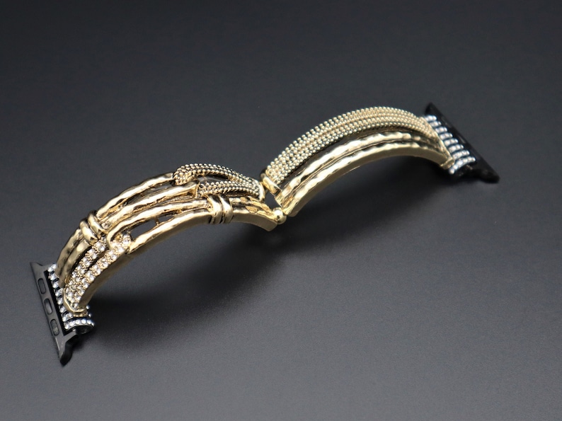 May include: Gold-tone Apple Watch band with a textured, hammered metal design and clear crystal accents. The band is designed to fit Apple Watch models.