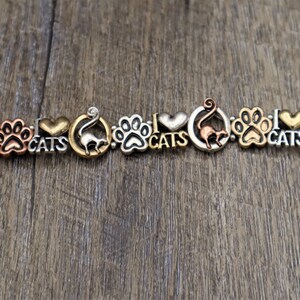 May include: A charm bracelet with a gold, silver, and copper finish. The bracelet features charms with paw prints, hearts, and cats. The charms are attached to a chain with a clasp. The bracelet is on a wooden surface.