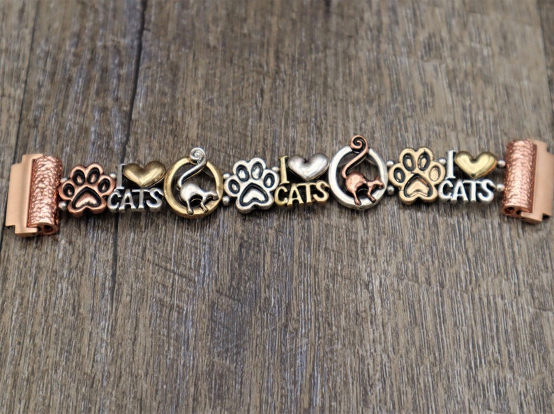 May include: A charm bracelet with a gold, silver, and copper finish. The bracelet features charms with paw prints, hearts, and cats. The charms are attached to a chain with a clasp. The bracelet is on a wooden surface.