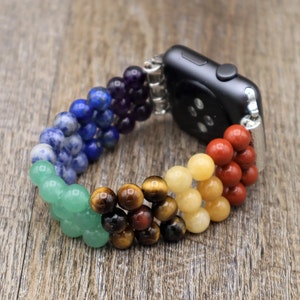 Op de afbeelding: Een regenboogkleurig kralenbandje voor de Apple Watch met een zwarte Apple Watch-wijzerplaat. Het bandje is gemaakt van verschillende soorten edelstenen, waaronder amethist, lapis lazuli, sodaliet, aventurijn, tijgeroog, citrien en rode jaspis.