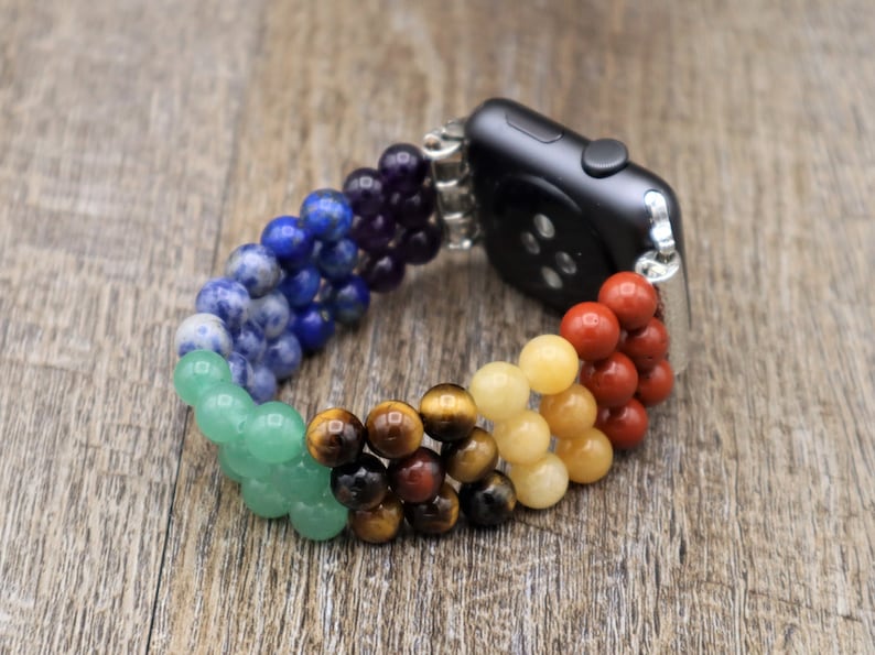 Op de afbeelding: Een regenboogkleurig kralenbandje voor de Apple Watch met een zwarte Apple Watch-wijzerplaat. Het bandje is gemaakt van verschillende soorten edelstenen, waaronder amethist, lapis lazuli, sodaliet, aventurijn, tijgeroog, citrien en rode jaspis.
