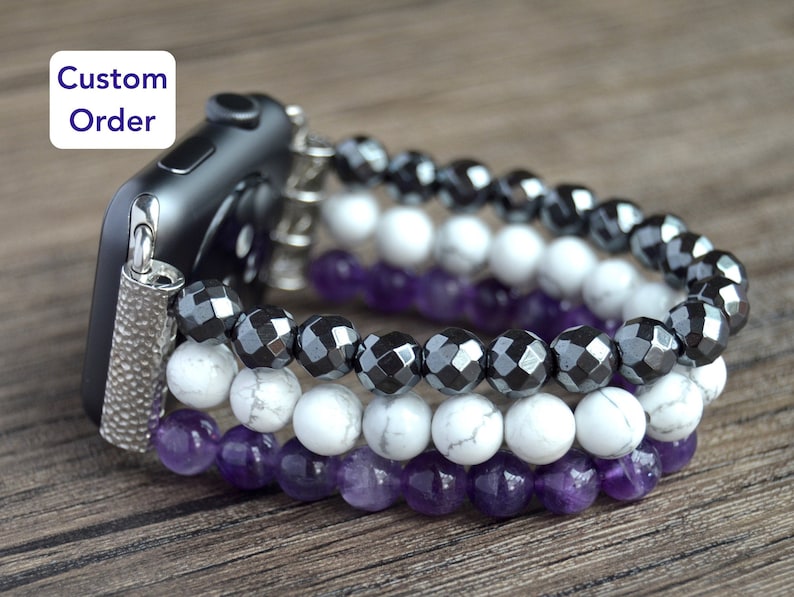 May include: A three-strand beaded Apple Watch band with black, white, and purple beads. The band is made of hematite, white howlite, and amethyst beads.