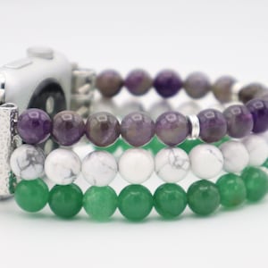 Puede incluir: Un Apple Watch plateado con una banda de cuentas de tres hilos. La banda está hecha de cuentas de amatista, howlite blanca y aventurina verde.