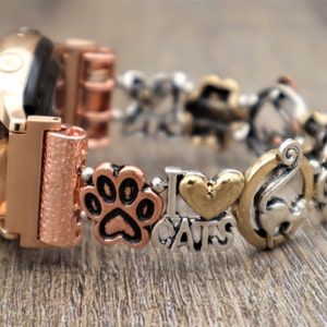 May include: A rose gold watch band with charms. The charms include a paw print, a heart with "I love cats" written on it, and a cat silhouette.