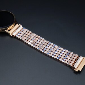 May include: A rose gold smartwatch band with a row of small, clear rhinestones. The band is made of metal and has a polished finish.