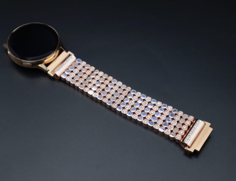 May include: A rose gold smartwatch band with a row of small, clear rhinestones. The band is made of metal and has a polished finish.
