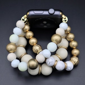 Puede incluir: Una pulsera de reloj de cuentas doradas, blancas y azul claro con una esfera de reloj Apple negra.