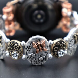 Pode incluir: Um bracelete de rel&oacute;gio prateado com um amuleto de borboleta de cobre e detalhes florais. O bracelete &eacute; feito de contas de metal e tem um mostrador de rel&oacute;gio prateado.