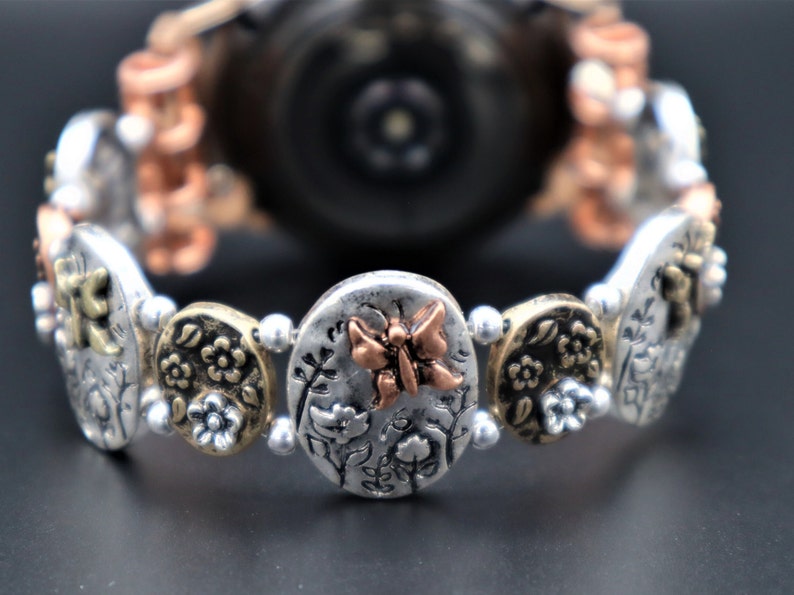 Pode incluir: Um bracelete de rel&oacute;gio prateado com um amuleto de borboleta de cobre e detalhes florais. O bracelete &eacute; feito de contas de metal e tem um mostrador de rel&oacute;gio prateado.