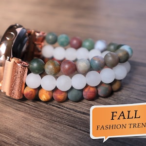 Puede incluir: Un smartwatch de oro rosa con una banda de cuentas multicolores. Las cuentas son en tonos de marrón, verde, blanco y rojo. La banda está hecha de piedras naturales. El texto FALL FASHION TRENDS está en un bocadillo de diálogo.