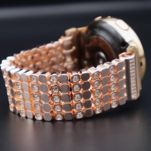 May include: A rose gold smartwatch band with a diamond-like pattern. The band is made of metal and has a polished finish.
