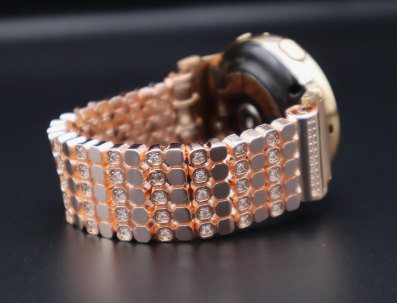 May include: A rose gold smartwatch band with a diamond-like pattern. The band is made of metal and has a polished finish.