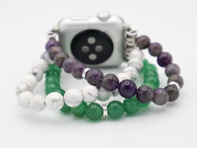 May include: A silver Apple Watch with a white, green, and purple beaded band. The band is made of three separate strands of beads.