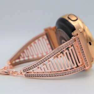 May include: A rose gold smartwatch band with a geometric design and clear rhinestones. The band is designed to fit a round smartwatch.