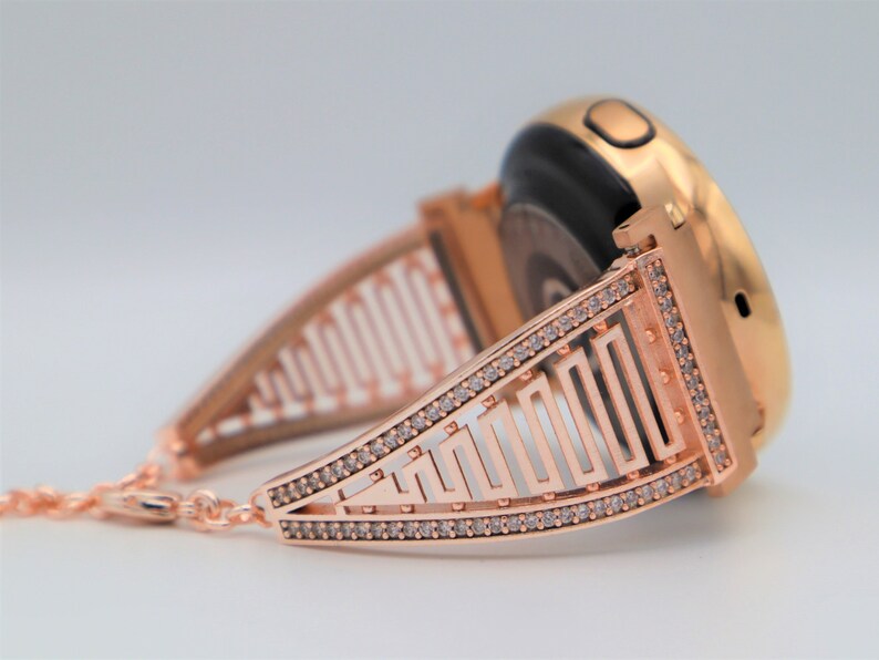 May include: A rose gold smartwatch band with a geometric design and clear rhinestones. The band is designed to fit a round smartwatch.