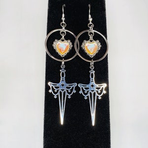 May include: A pair of silver earrings with a heart-shaped charm and a dagger-shaped pendant. The heart charm has an iridescent finish with orange, pink, and blue hues. The dagger pendant is detailed with intricate designs.